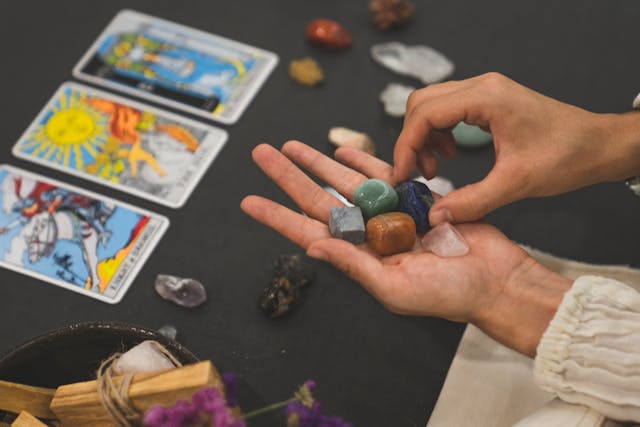 Mano sosteniendo cristales de diferentes colores frente a una mesa con cartas de tarot y palo santo, en una tienda esotérica de Madrid, ideal para quienes buscan lecturas y sanación energética.